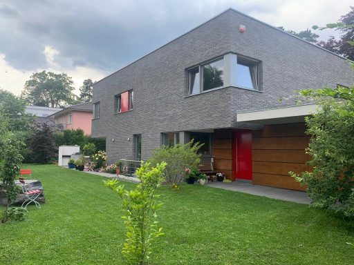 Modernes Haus mit grauer Fassade, roter Tür und angelegtem Garten.
